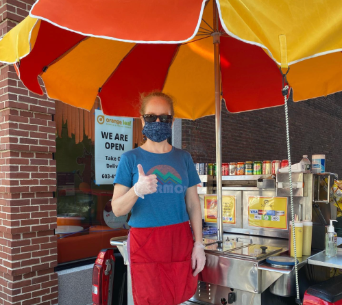 A vendor in a mask and gloves gives a thumbs up at a food cart under a colorful umbrella, with a sign that says "We Are Open."