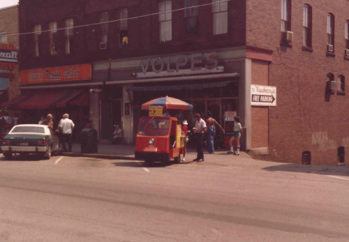 A colorful food cart outside a brick building with "Volpe's" sign, surrounded by people and parked cars.