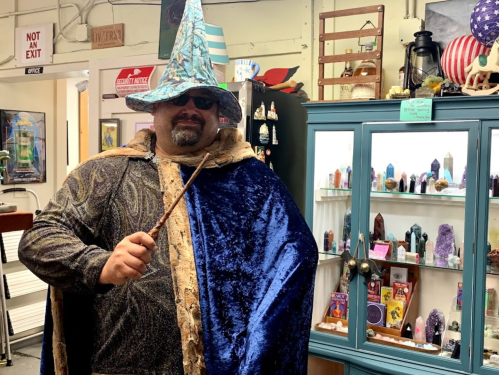 A person in a wizard costume with a blue hat and cape stands in a shop filled with crystals and mystical items.