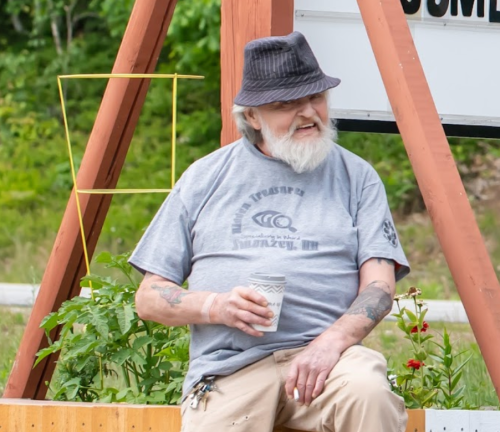 An older man with a beard sits on a bench, holding a coffee cup, wearing a gray shirt and a black hat.
