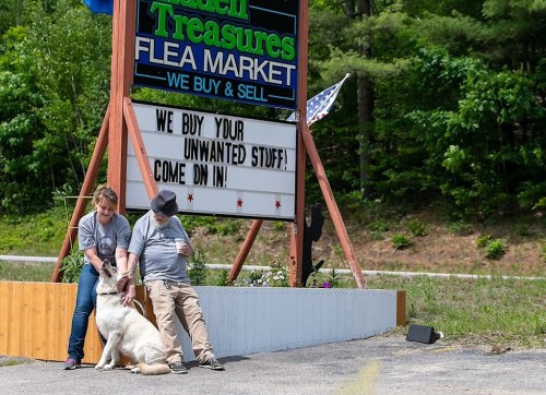 A woman and a man sit outside a flea market, petting a dog near a sign that says "We buy your unwanted stuff!"