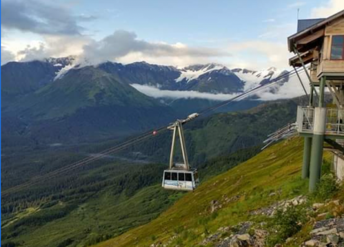 A gondola ascends a mountain, surrounded by lush greenery and snow-capped peaks under a cloudy sky.