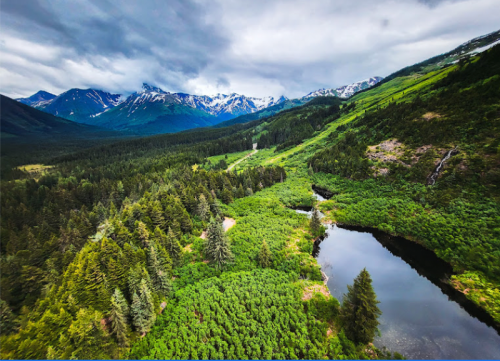 Aerial view of a lush green landscape with a river, mountains, and cloudy skies in the background.