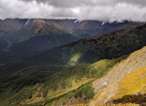 A scenic mountain landscape with lush forests, rocky slopes, and clouds hovering over the peaks.