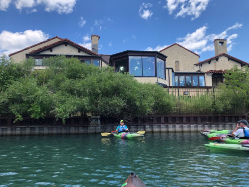 Two kayakers on a calm waterway near a large house surrounded by greenery and blue skies.