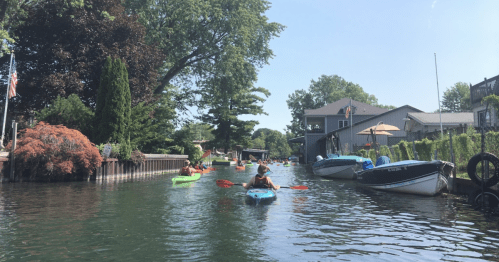 A serene waterway with kayakers paddling among boats and lush greenery on a sunny day.