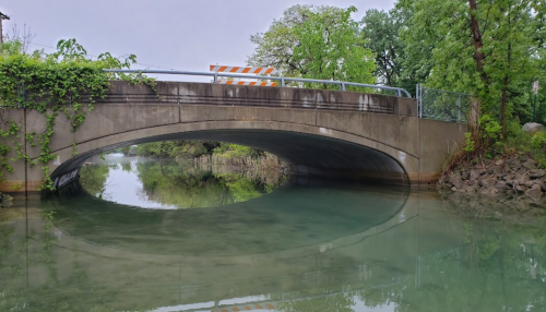 A concrete bridge arches over a calm, greenish river, surrounded by lush trees and vegetation.