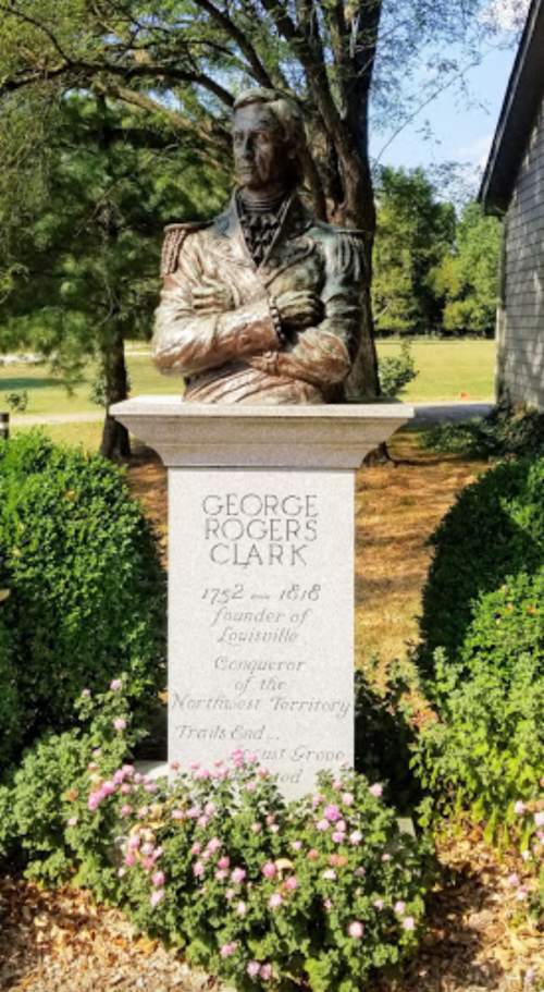 Statue of George Rogers Clark, founder of Louisville, with a stone pedestal and surrounding greenery and flowers.
