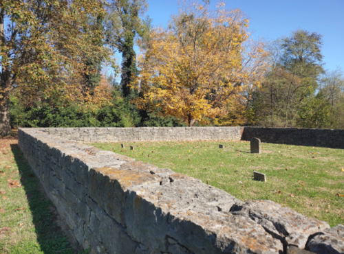 A stone wall encloses a grassy area with scattered small stones, surrounded by trees with autumn foliage.
