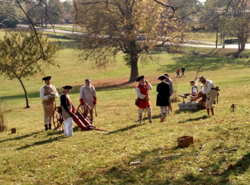 A group of people in historical costumes reenacting a scene outdoors, with trees and a road in the background.