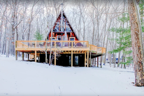A snowy landscape featuring a large A-frame cabin with a wooden deck surrounded by trees.