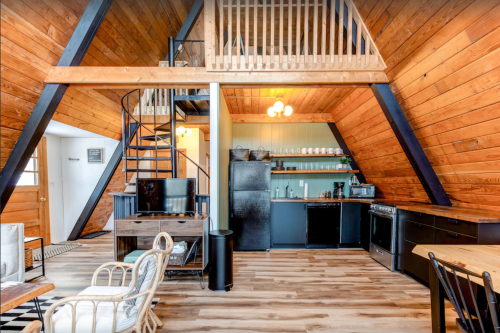 Cozy A-frame interior with wooden walls, spiral staircase, kitchen area, and seating space. Bright and inviting atmosphere.
