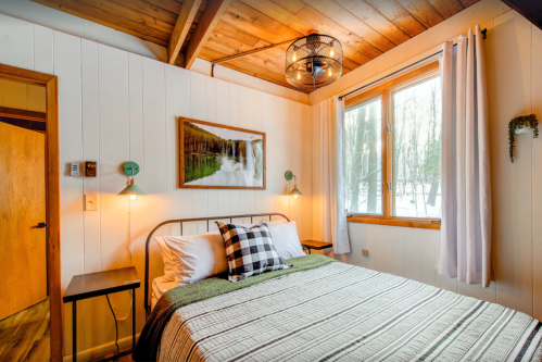 Cozy bedroom with a wooden ceiling, a bed with plaid accents, and a large window overlooking a snowy landscape.
