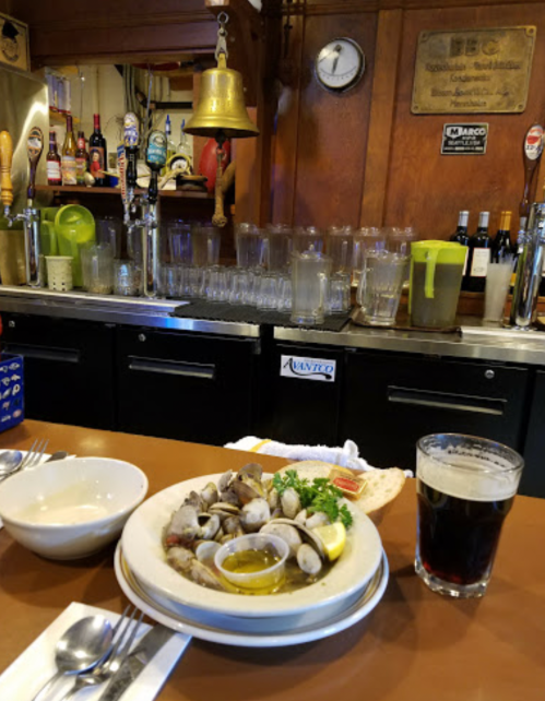 A plate of seafood with lemon and herbs, a glass of dark beer, and a bar with drinks in the background.