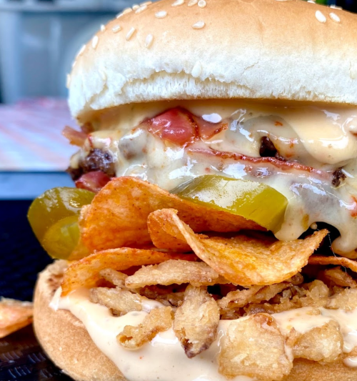 Close-up of a sesame seed burger with layers of meat, cheese, tomatoes, pickles, and crispy potato chips.