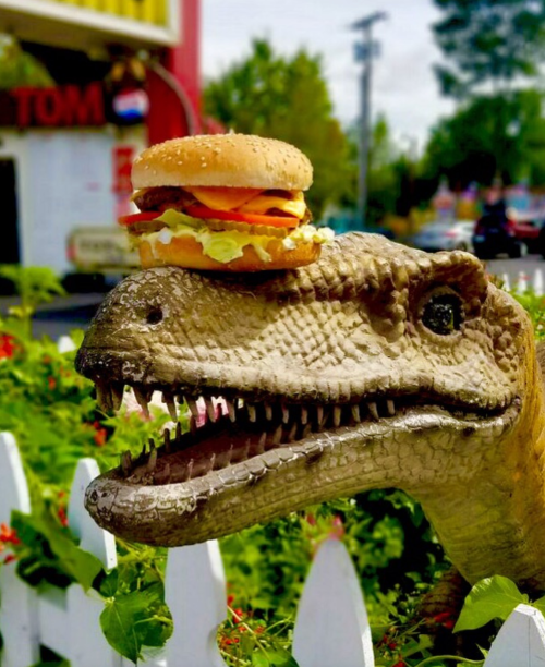A dinosaur statue with a hamburger on its head, surrounded by greenery and a colorful background.