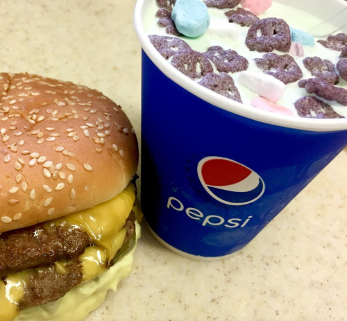 A sesame seed burger with cheese and a cup of Pepsi topped with colorful marshmallows and cereal.