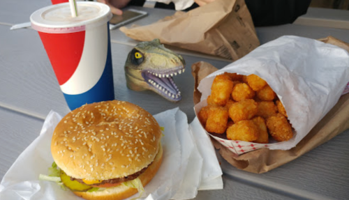 A sesame seed burger, tater tots in a bag, a drink, and a toy dinosaur on a table.