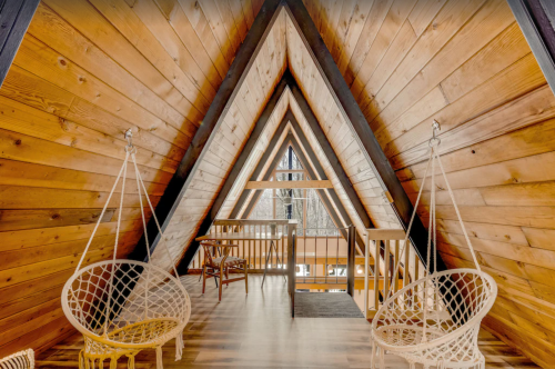 Cozy attic space with wooden walls, featuring hanging chairs and a view of a snowy landscape through large windows.