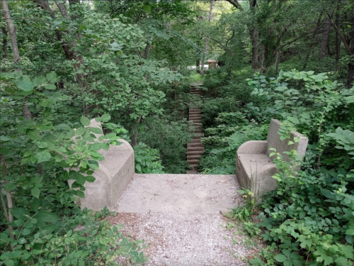 A stone overlook surrounded by lush greenery, leading to a staircase descending into a wooded area.