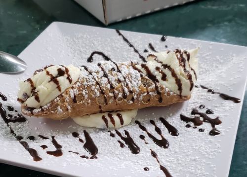 A delicious cannoli filled with cream, drizzled with chocolate sauce, and dusted with powdered sugar on a white plate.