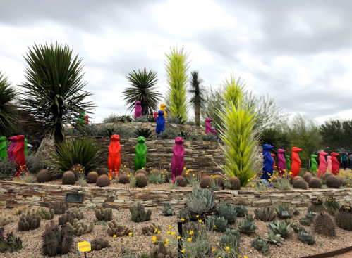 Colorful sculptures in vibrant hues arranged on a stone mound surrounded by desert plants under a cloudy sky.