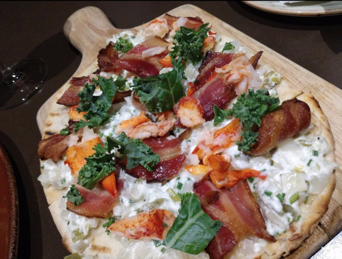 A wooden board holds a pizza topped with bacon, shrimp, greens, and creamy sauce, showcasing a delicious combination.
