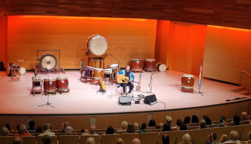 A musician performs on stage with various drums and instruments, while an audience watches attentively.