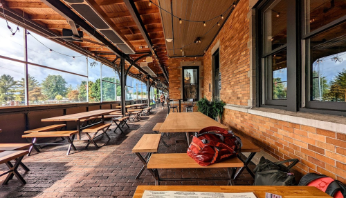 A spacious outdoor dining area with wooden tables, benches, and a brick wall, surrounded by greenery and string lights.