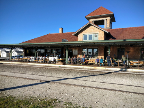 A historic train station with outdoor seating, filled with people enjoying a sunny day.