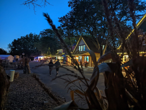 A twilight scene of a charming street lined with lit-up buildings and autumn decorations, with two people walking.