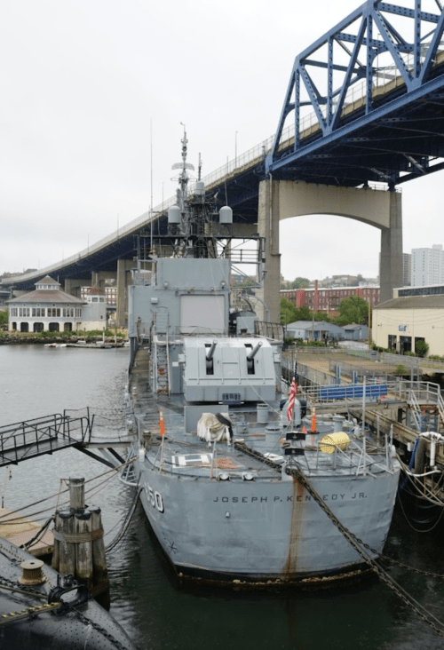 A naval ship docked near a bridge, with buildings and a cloudy sky in the background.