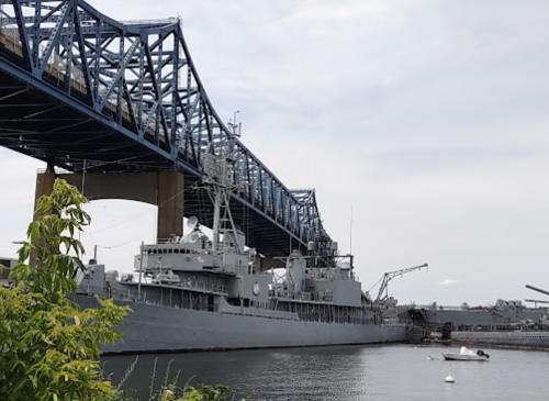 A large gray ship docked under a blue bridge, with calm water and greenery in the foreground.