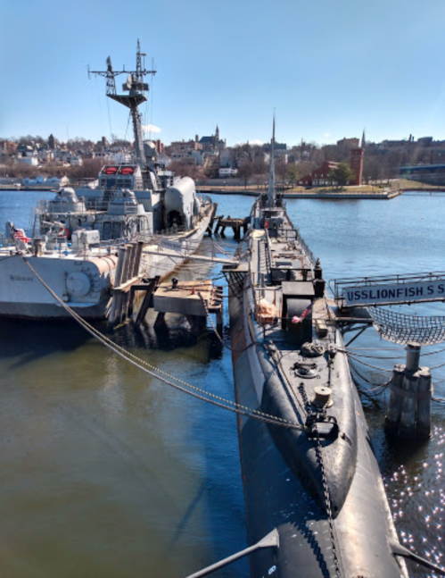 A submarine and a naval ship docked at a waterfront, with a clear blue sky and cityscape in the background.