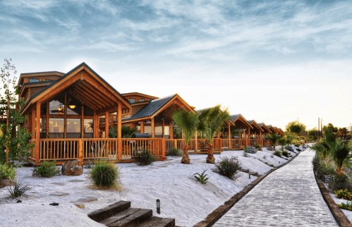 A row of wooden cabins with porches, surrounded by palm trees and a stone pathway under a blue sky.