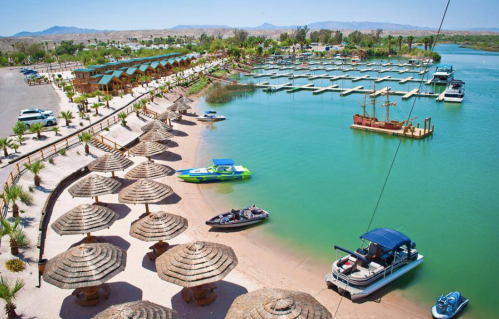 A scenic view of a marina with boats, sandy beach, palm trees, and thatched umbrellas under a clear blue sky.