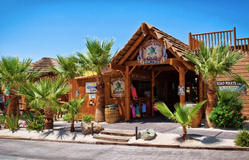 A tropical-themed restaurant entrance with palm trees and a wooden structure under a clear blue sky.