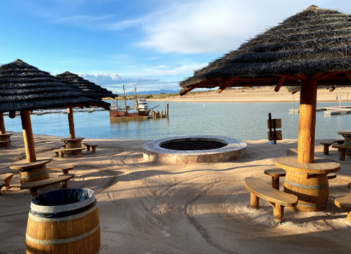 A sandy area with thatched-roof huts, wooden tables, and a fire pit by a calm lake with a boat in the background.