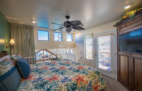 Cozy bedroom with floral bedding, large windows, a ceiling fan, and a TV, featuring a view of a deck outside.