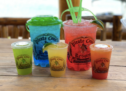 Five colorful drinks in cups and buckets, featuring the "Pirate Cove Resort" logo, on a wooden table.