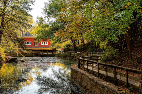 A serene scene of a winding path by a calm waterway, surrounded by trees in autumn colors and a red building in the background.