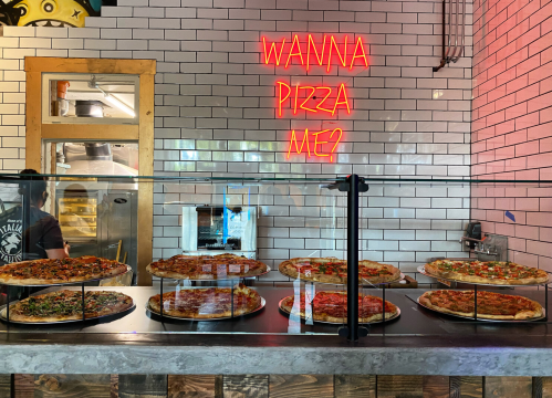 A display of various pizzas in a restaurant, with a neon sign that says "Wanna pizza me?" in the background.