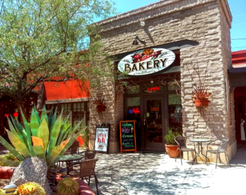 A charming bakery with a stone facade, outdoor seating, and vibrant plants in front, inviting customers inside.