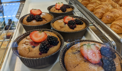 Five muffins topped with strawberries and blackberries, displayed in a bakery case alongside croissants.