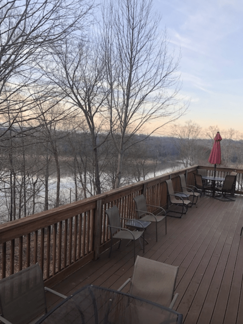 A wooden deck with chairs overlooks a river and bare trees under a pastel sky at dusk.