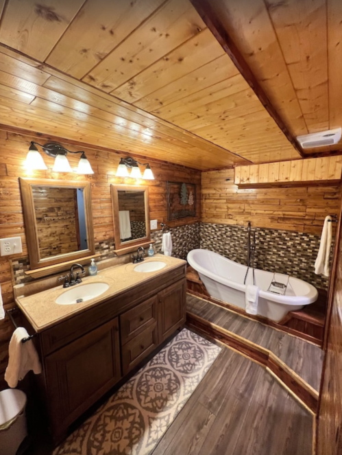 Cozy wooden bathroom featuring a double sink, mirrors, and a freestanding bathtub with a patterned rug.