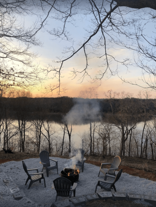 A serene lakeside scene at sunset, featuring a fire pit surrounded by chairs and trees reflecting on the water.