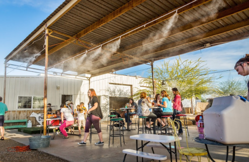 A lively outdoor gathering under a shaded structure, with people socializing and misting for cooling.