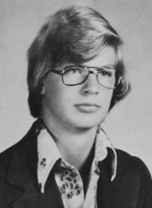 A young man with glasses and a collared shirt poses for a school portrait, wearing a dark blazer.
