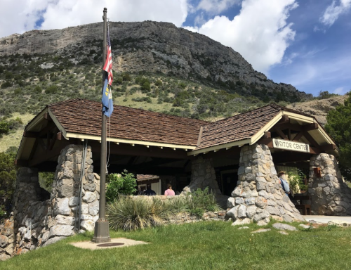 A stone visitor center with a flagpole, set against a mountainous backdrop and blue sky with clouds.
